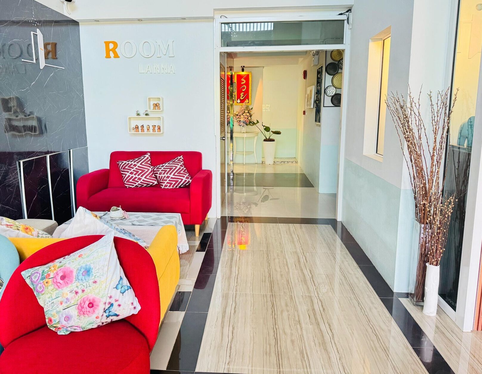 Un hall de hotel con un sofá rojo, un sofá amarillo y un televisor con marco negro. Hay un cartel que dice "ROOM LANTA" y una mesita con un jarrón de flores.