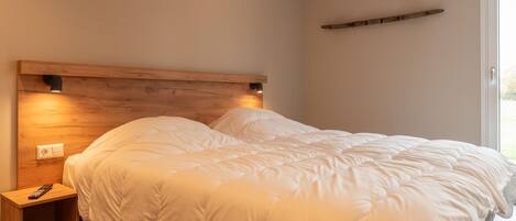 A neatly made bed with a wooden headboard, a bedside table with a remote control, and a small shelf above the bed.