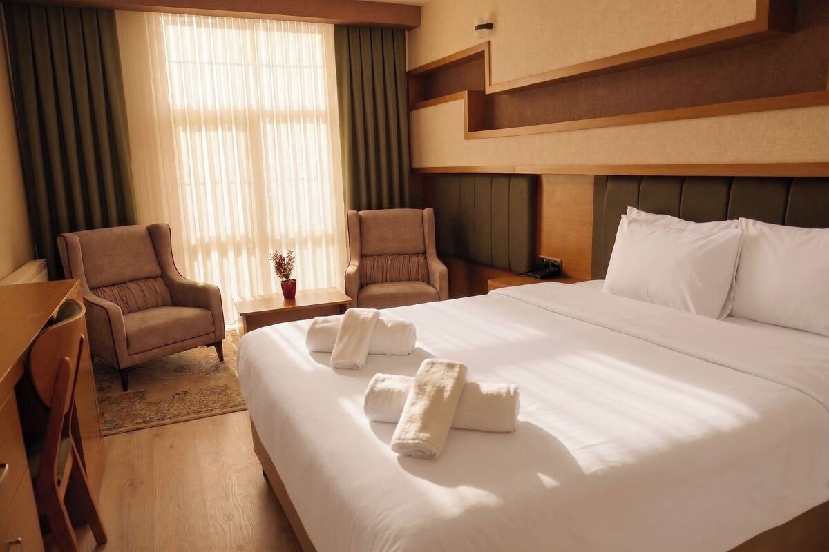 Una habitación de hotel con una cama grande, dos sillones, un escritorio y un ventanal con cortinas.