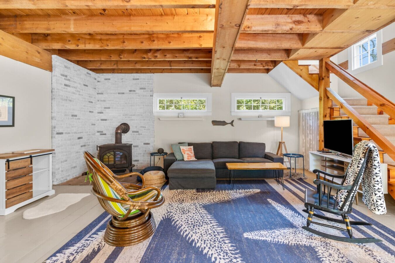A cozy living room with a wooden ceiling, a fireplace, a sofa, a rocking chair, and a staircase.