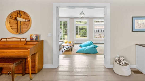 A room with a wooden piano, a round mirror, a white door leading to a balcony with a view of greenery, and a hanging light fixture. - Gorgeous Home 1 mile to Beach, by Cape Cod Bikepath (Dennis)