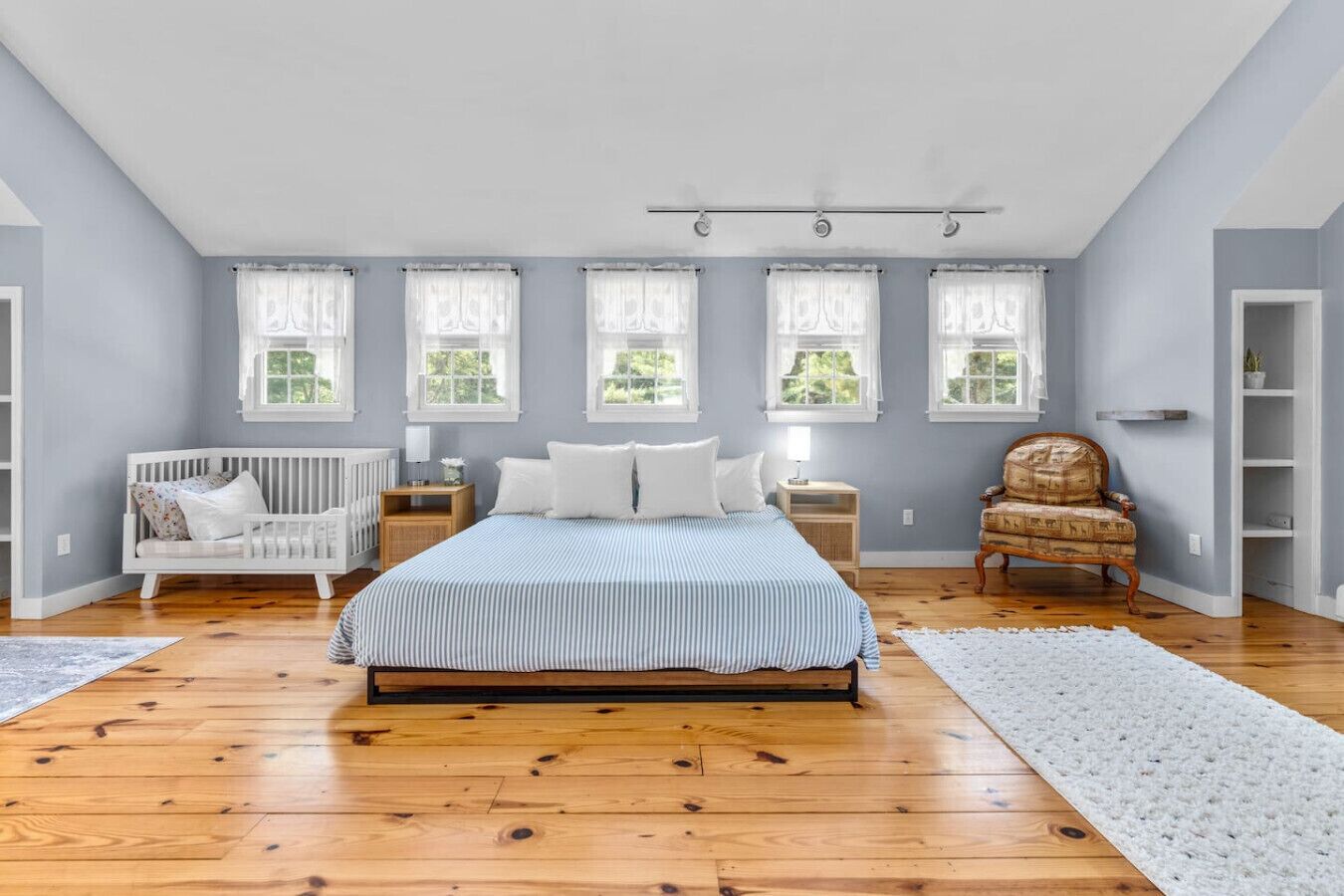 Ein Schlafzimmer mit einem großen Bett, Holzboden, einem weißen Babybett, einem Schaukelstuhl und einem Fenster mit Vorhängen.