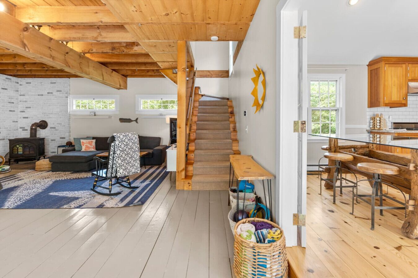 A cozy cabin interior with a wooden staircase, a living area with a sofa and a fireplace, and a kitchen with wooden bar stools.