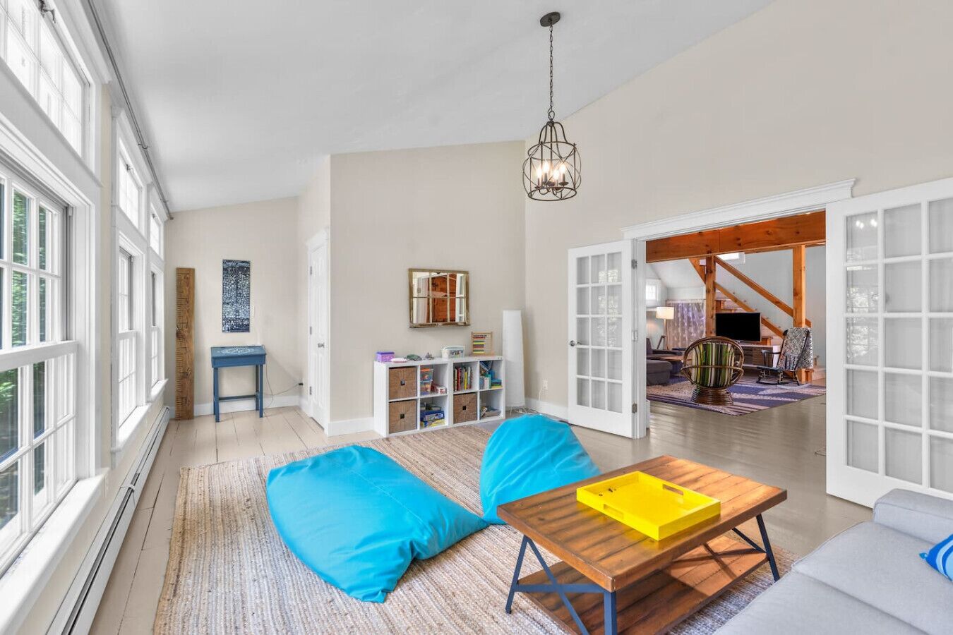 A spacious living room with large windows, a wooden table, blue cushions, and a bookshelf.
