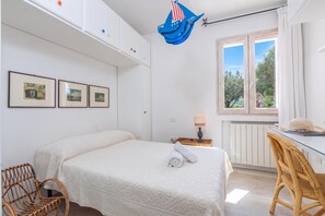 A bedroom with a bed, a desk with a lamp, a wicker chair, and a window with a view of trees.