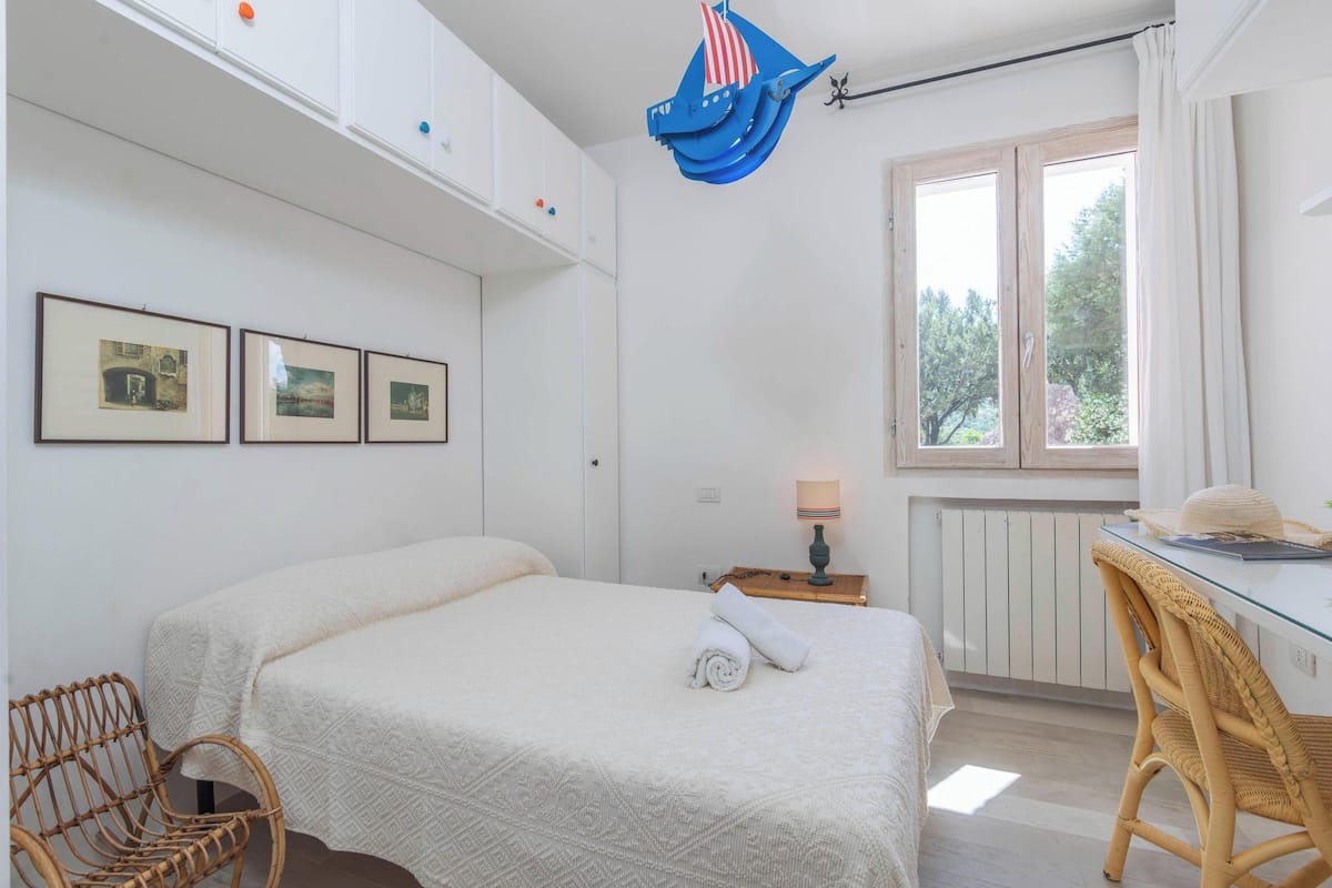 A bedroom with a bed, a desk, a chair, and a window with a view of trees.