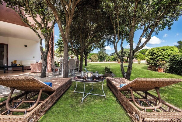 An outdoor seating area with wicker lounge chairs and a table, surrounded by trees and a well-maintained lawn.