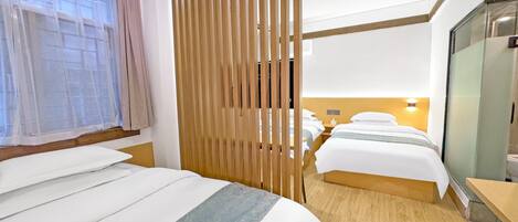 Ett hotellrum med två sängar, en avskärmande vägg i trä, en duschkabin med glasväggar och ett fönster med tunna, genomskinliga gardiner.