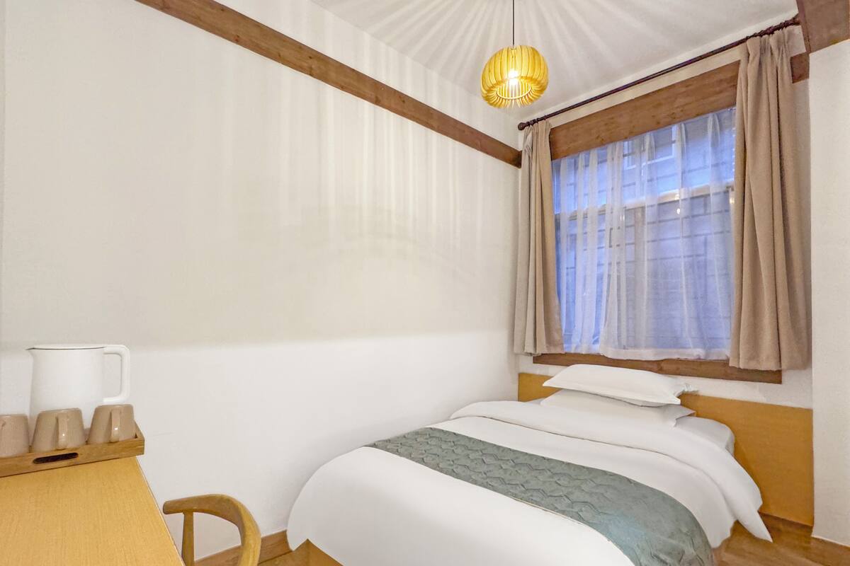 A room with a bed, a wooden desk with a cup holder, a chair, and a window with curtains.