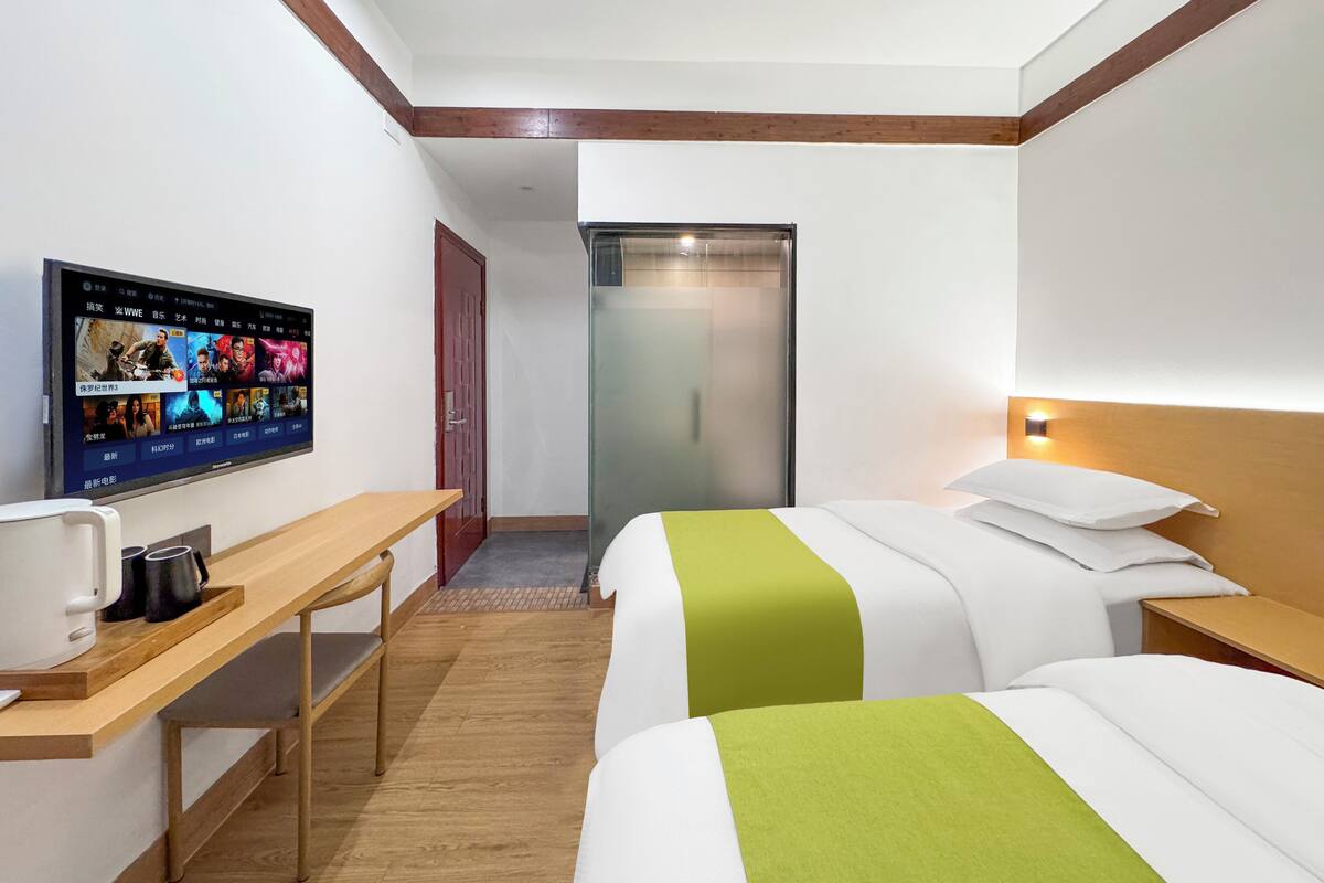 A hotel room with a TV on the wall, a desk with a coffee maker, two beds with green and white bedding, and a wooden headboard.