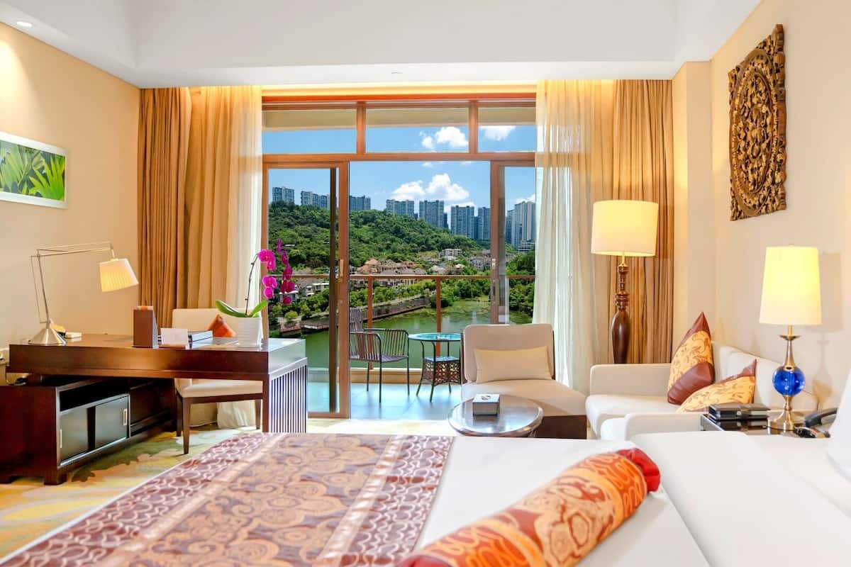 A hotel room with a large bed, a desk with a lamp, a chair, a small table, and a view of a cityscape and greenery through large windows.