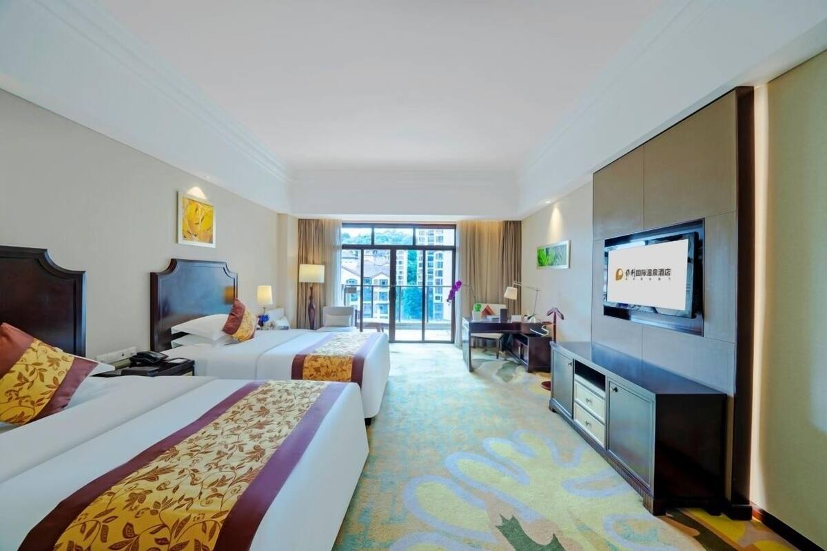 A hotel room with two beds, a desk, a television, and a view of the outside.