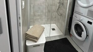 Un bagno compatto con doccia vetrata, una lavatrice e asciugatrice affiancate e una piccola panca con un asciugamano.