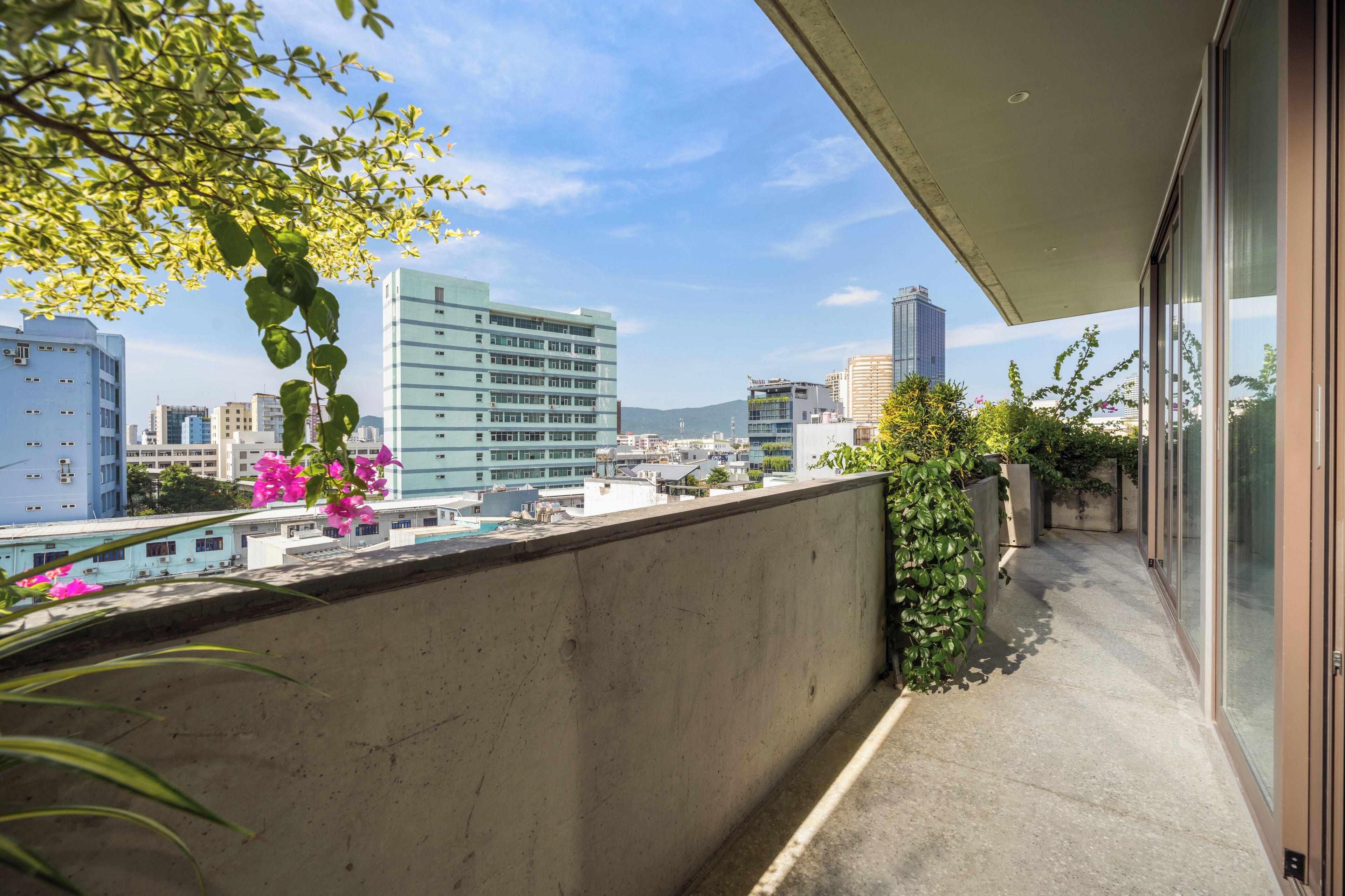 A balcony with a concrete wall, a view of city buildings, and some greenery.