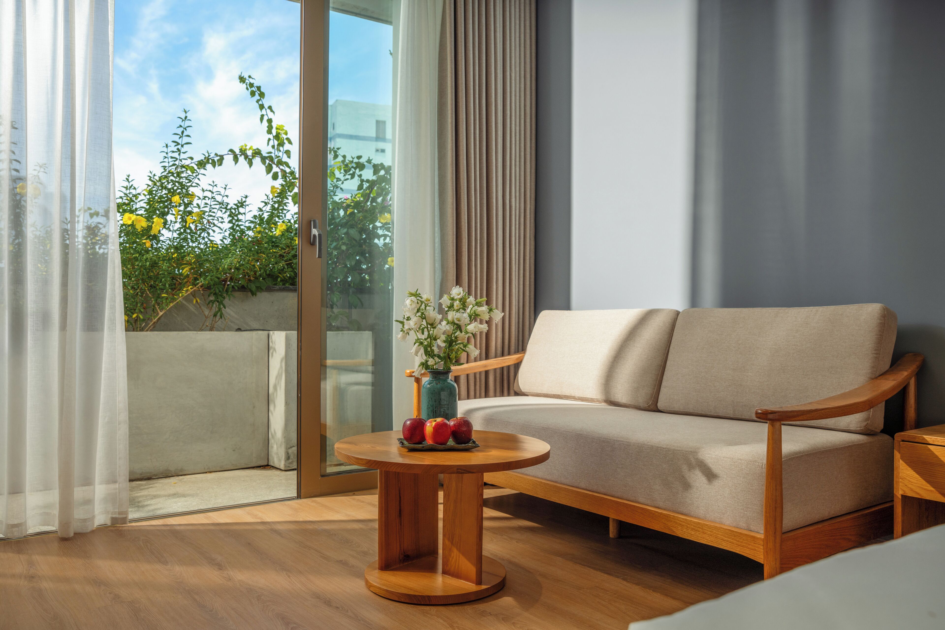 A modern living room with a sofa, a round wooden coffee table with fruits and flowers, and a view of greenery through a glass door.