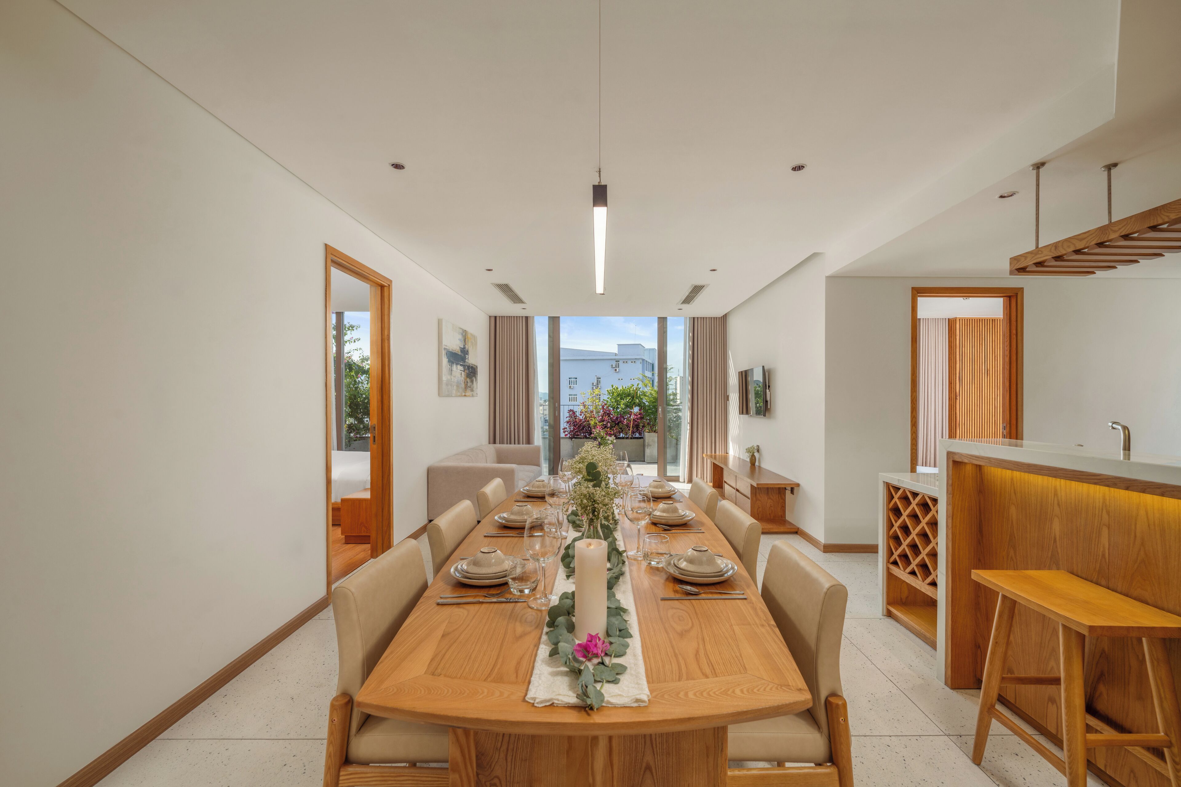 A modern dining area with a wooden table set for six, flanked by beige chairs. A tall white candle adorns the center of the table, and a vase with greenery and a pink flower is placed on the table's center.