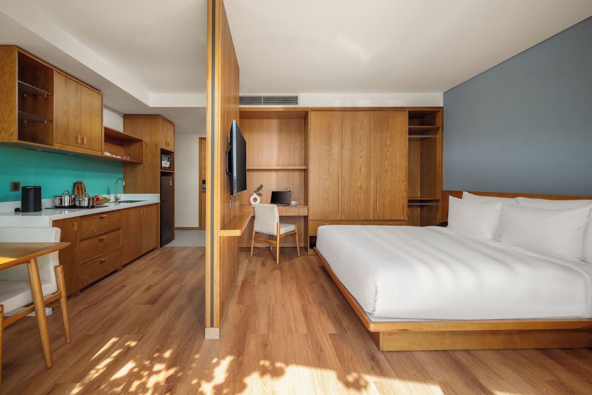 Une chambre d’hôtel moderne avec un plancher en bois, un lit, un bureau avec une chaise, un coin cuisine et un téléviseur.