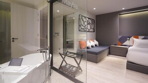 Une chambre d’hôtel moderne dotée d’une paroi en verre, d’une baignoire, d’un lit et d’un coin salon avec des coussins.