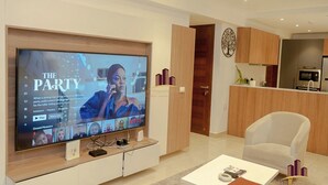 A modern living room with a large flat-screen TV displaying a show, a white sofa, a coffee table, and wooden cabinetry. - Solaris Cozy Haven (Accra)