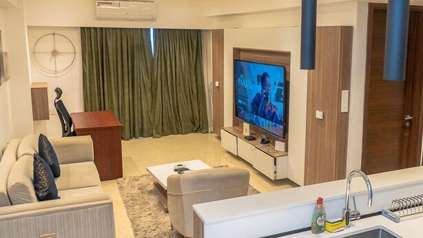 A modern living room with a TV, sofa, and coffee table. There is a kitchen area with a sink and a window. - Solaris Cozy Haven (Accra)