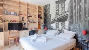 A hotel room with a bed, a desk with a computer, a bookshelf, and a large cityscape mural on the wall.
