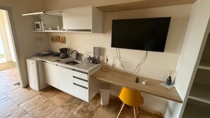 Ett kompakt kök med en inbyggd mikrovågsugn, en kaffebryggare och ett handfat. Det finns en liten papperskorg och en gul stol. En platt-TV är monterad på väggen ovanför bänkskivan.
