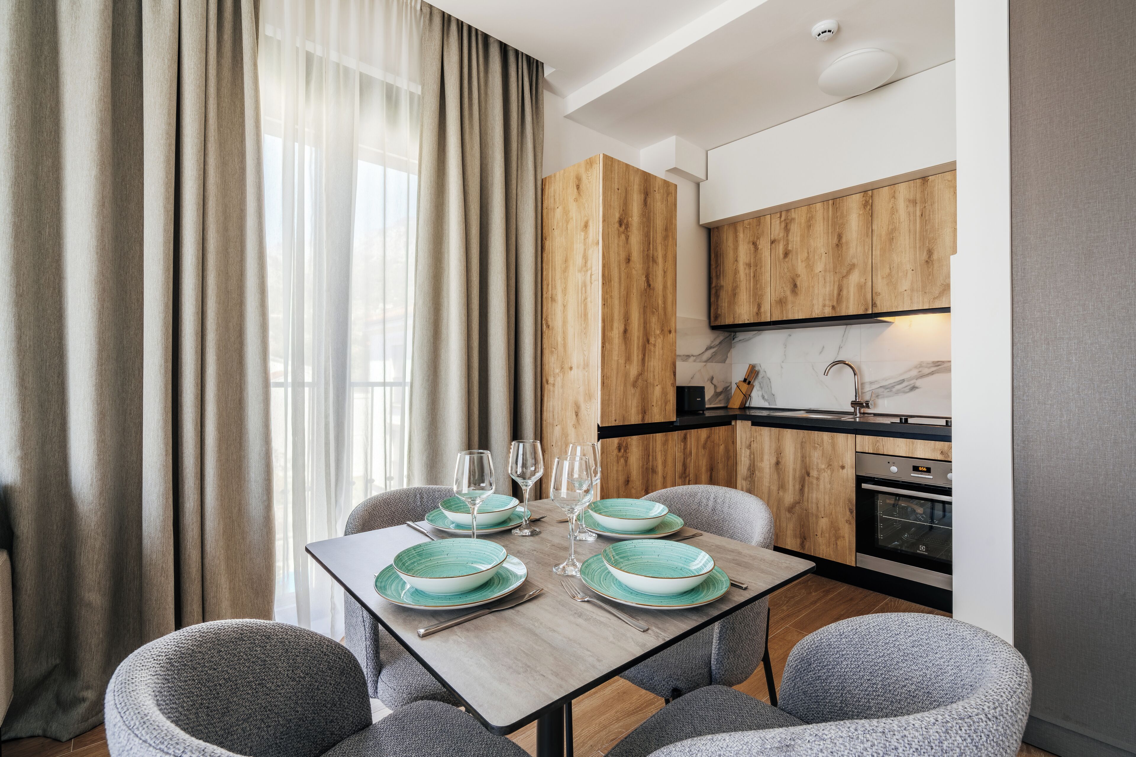 A modern dining area with a wooden table, grey upholstered chairs, and a kitchen in the background.