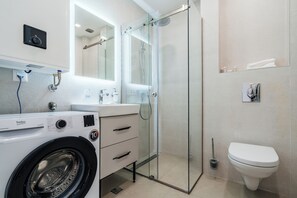 A modern bathroom with a glass-enclosed shower, a white toilet, a washing machine, and a sink with a mirror above it.