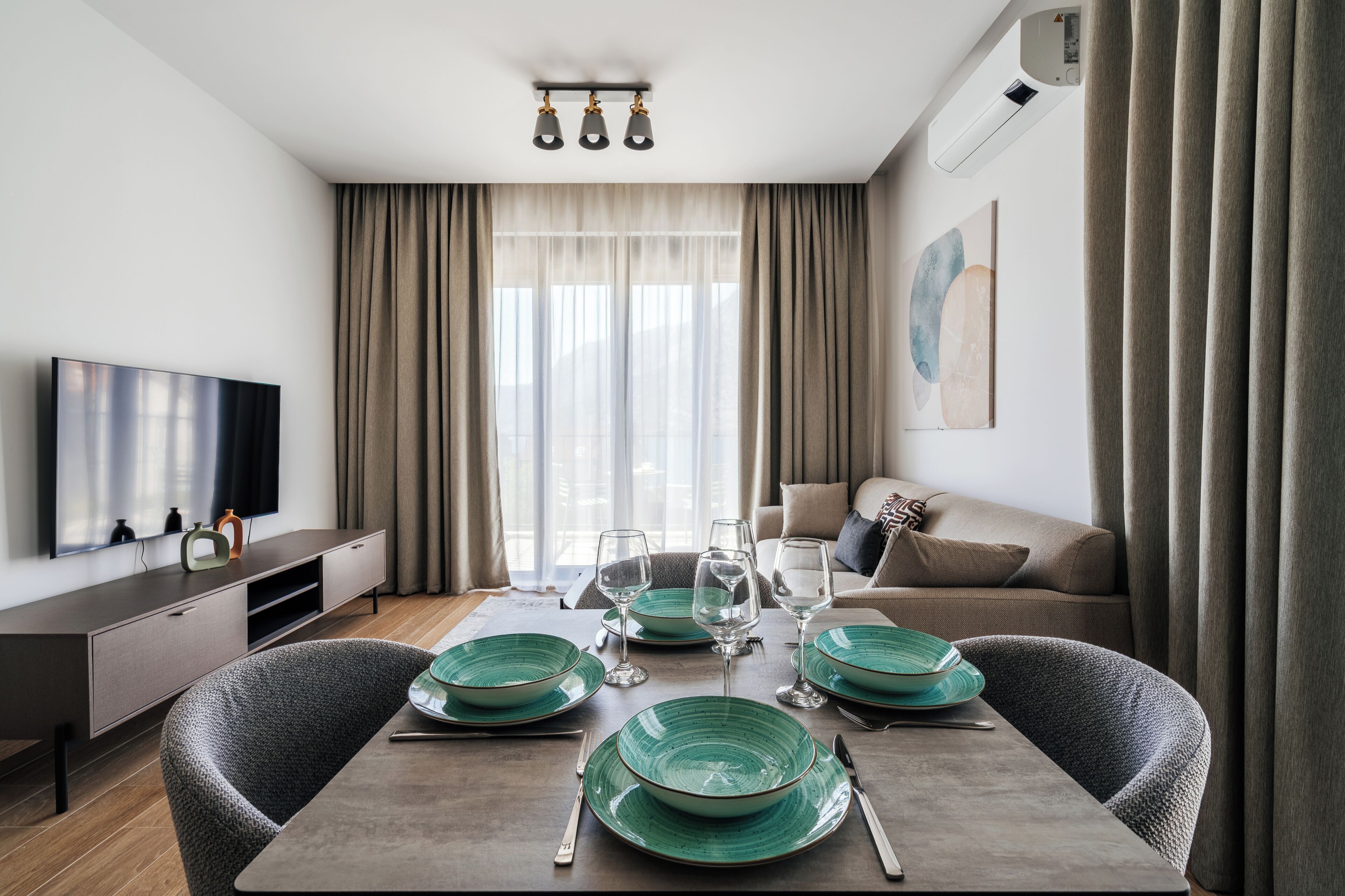 A modern living room with a dining table set for four, a flat-screen TV, a sofa, and a large window with curtains.