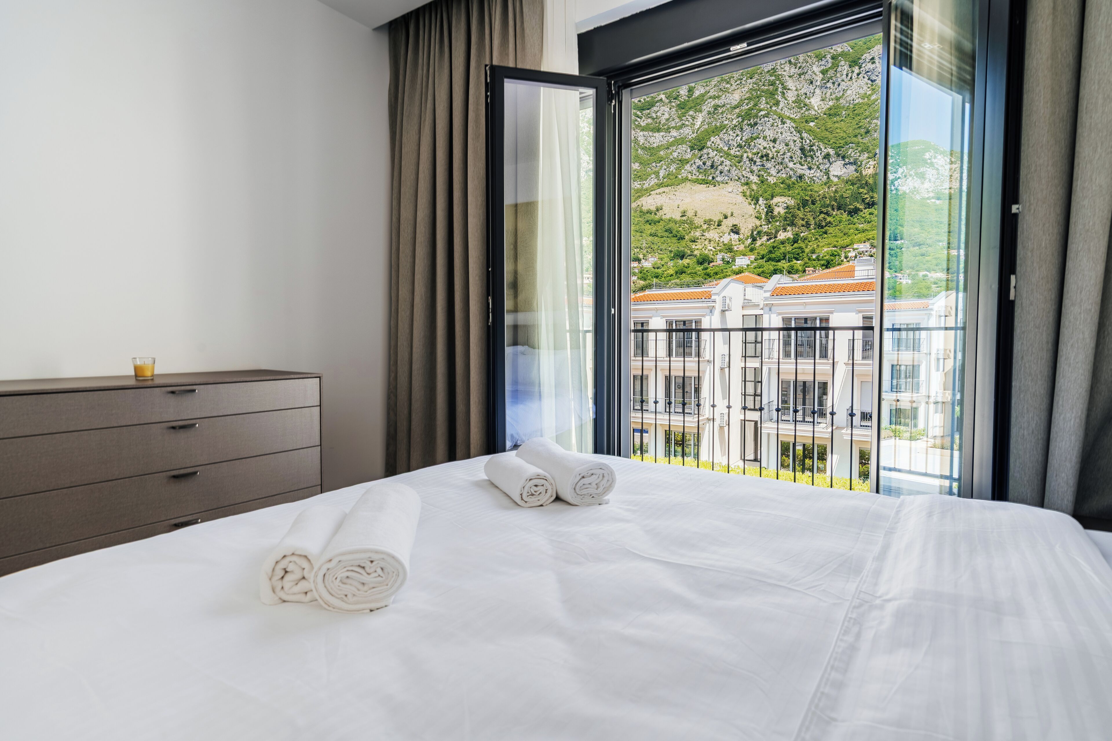 Een hotelkamer met een bed, een dressoir en een balkon met uitzicht op gebouwen en groen.