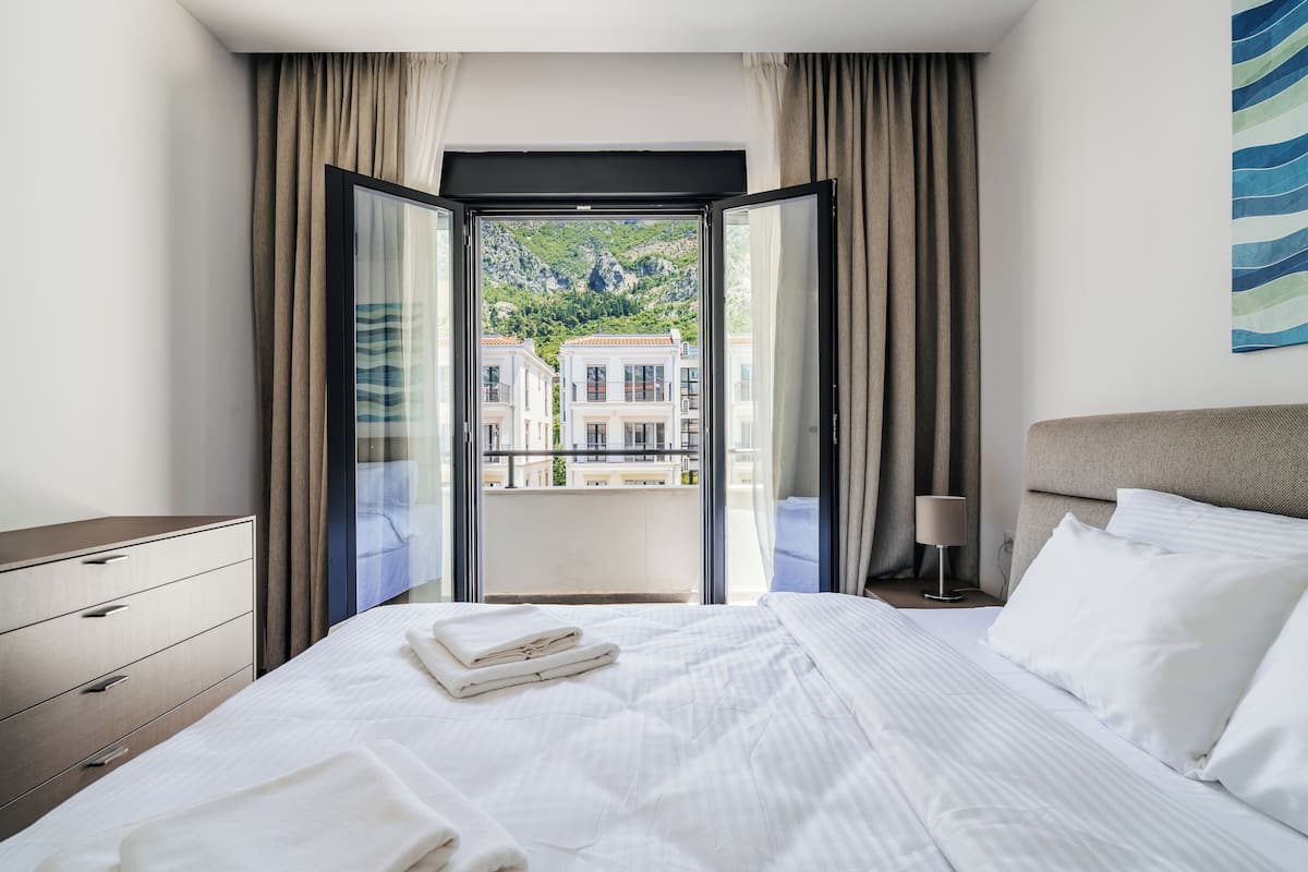 Une chambre d’hôtel avec un grand lit, une table de chevet, une lampe et une vue sur les bâtiments, visible à travers une porte ouverte.