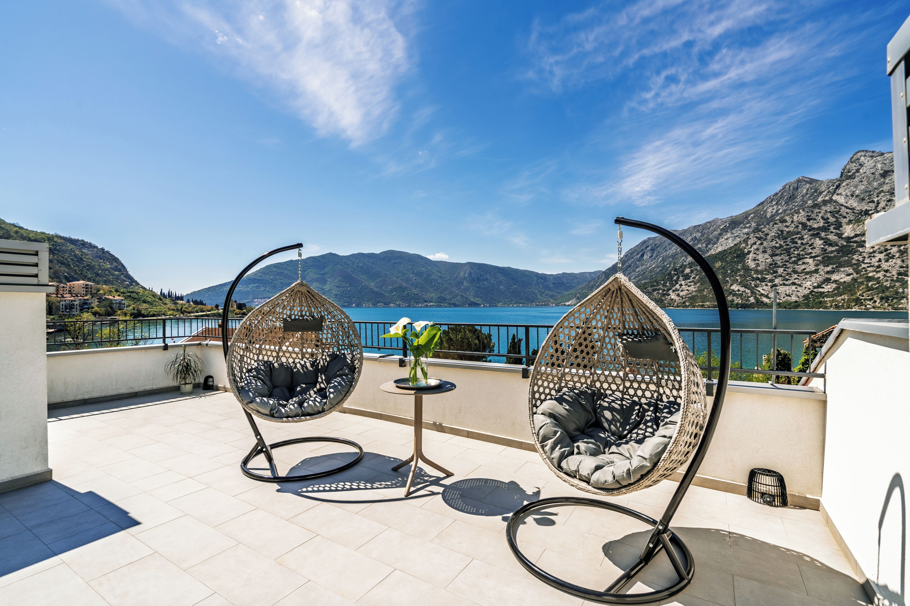 A terrace with two hanging chairs, a small table with a vase, and a view of mountains and a lake.