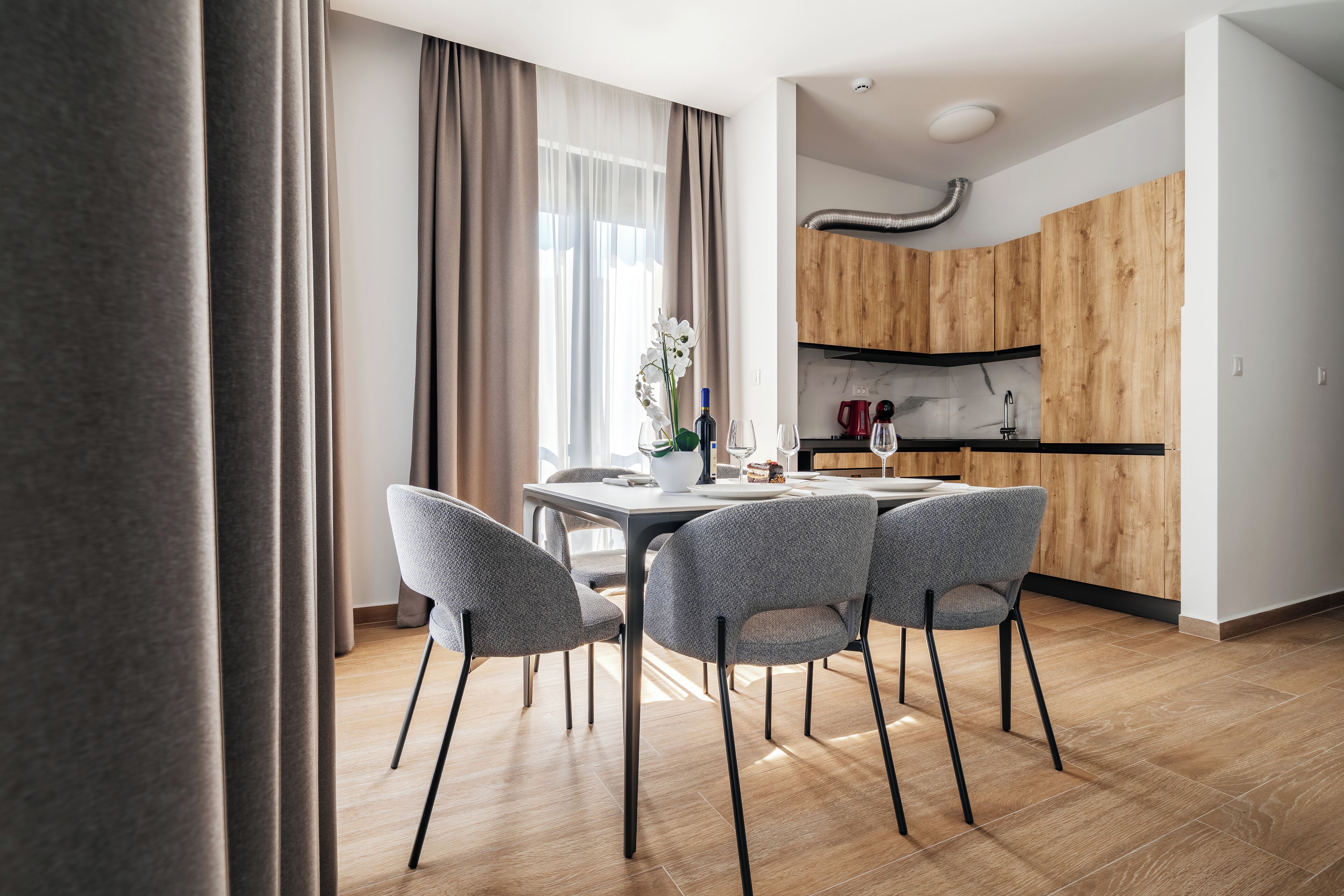 Un comedor moderno con una mesa y cuatro sillas, una orquídea en maceta y una cocina con alacenas de madera y salpicadero de mármol.