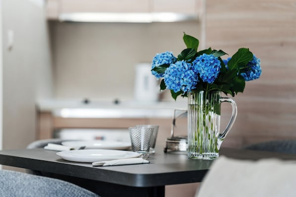 Una mesa de comedor con una jarra de vidrio transparente con flores azules, una taza de metal y platos blancos.