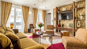 A living room with a sofa, armchair, and coffee table, a TV, and a bookshelf. - Merveil Luxury Suites - Rougemont I-II-III (Paris)