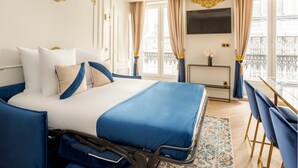 A hotel room with a large bed, a desk with a chair, a TV mounted on the wall, and a balcony with a railing. - Merveil Luxury Suites - Rougemont I-II-III (Paris)
