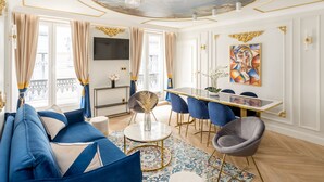 A spacious living room with a large sofa, a dining table with chairs, a TV mounted on the wall, and a ceiling with a mural. - Merveil Luxury Suites - Rougemont I-II-III (Paris)