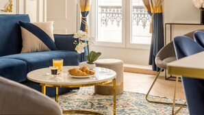 A modern living room with a blue sofa, a round coffee table with a gold base, and a small round ottoman. There is a glass of orange juice and a croissant on the table. - Merveil Luxury Suites - Rougemont I-II-III (Paris)