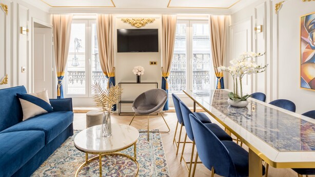 A modern living room with a blue sofa, a round coffee table, and a dining table with blue chairs. - Merveil Luxury Suites - Rougemont I-II-III (Paris)