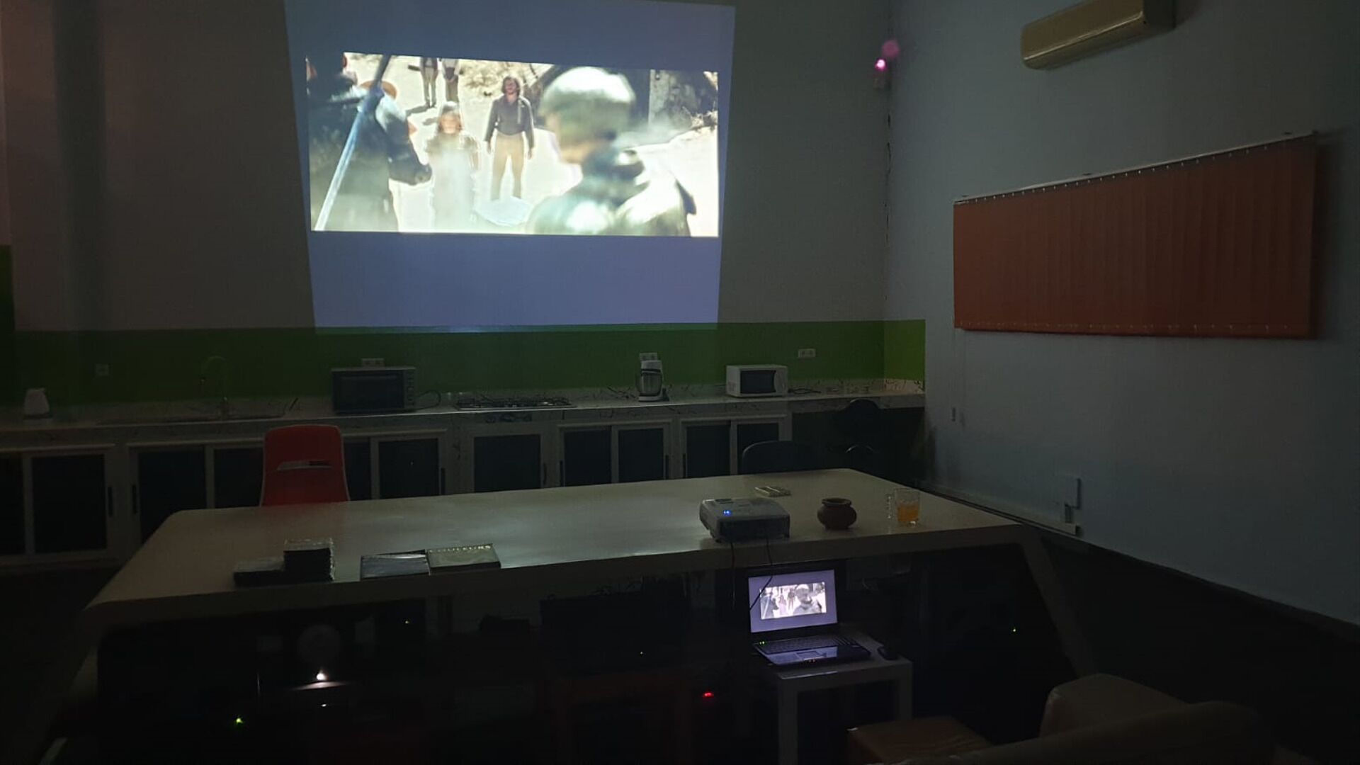 Un salón de conferencias con una pantalla grande mostrando un video, un proyector, una computadora portátil y una mesa pequeña con una taza y un vaso.