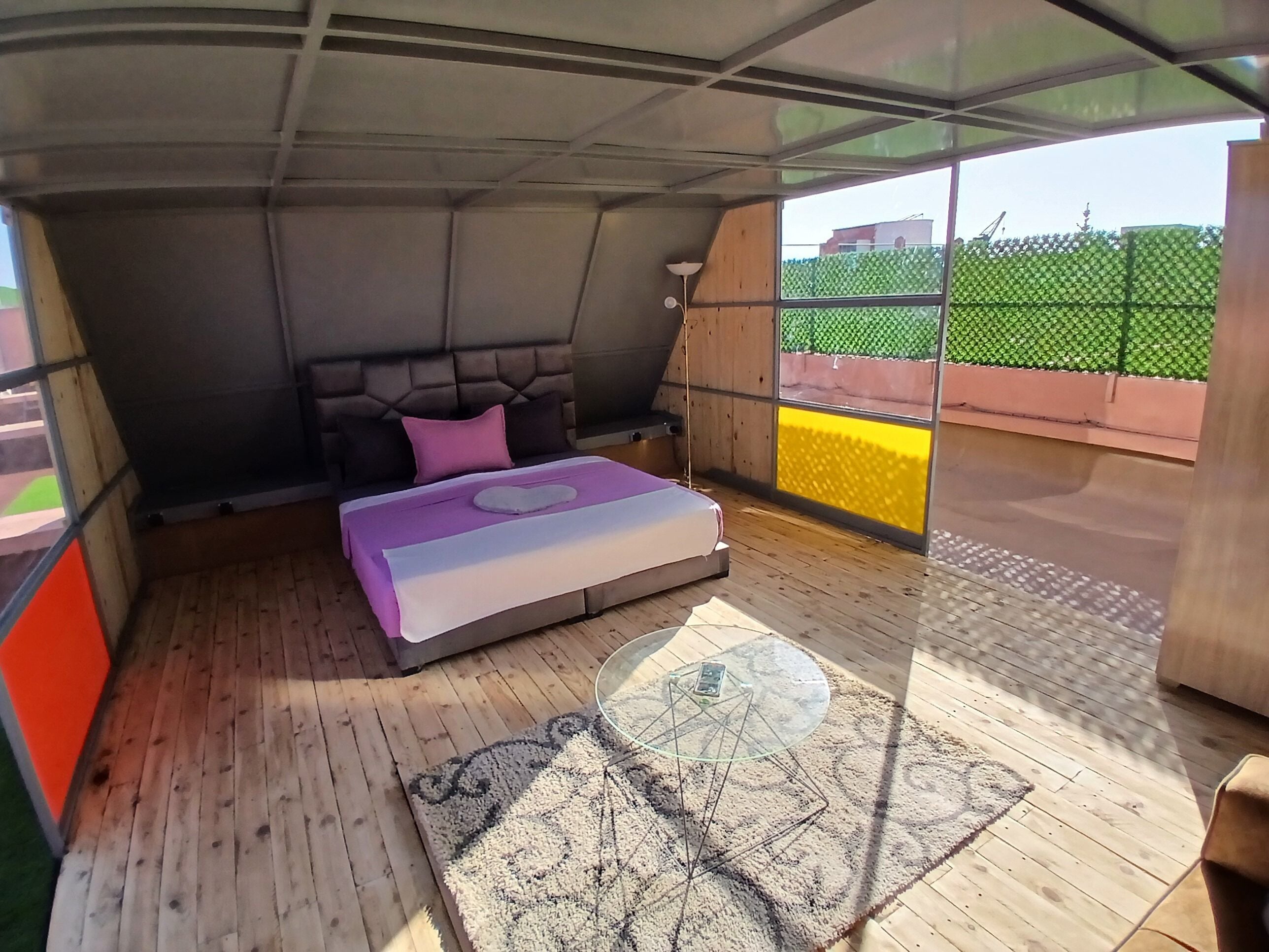 A bedroom with a bed, a glass table, and a view of the outside.