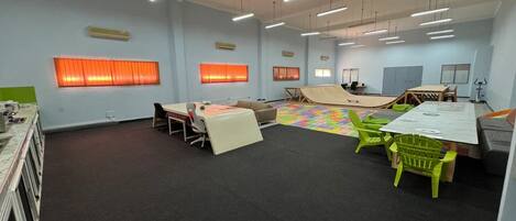 Una habitación amplia con una mesa grande, sillas, un pequeño escenario y un tapete de juego colorido.