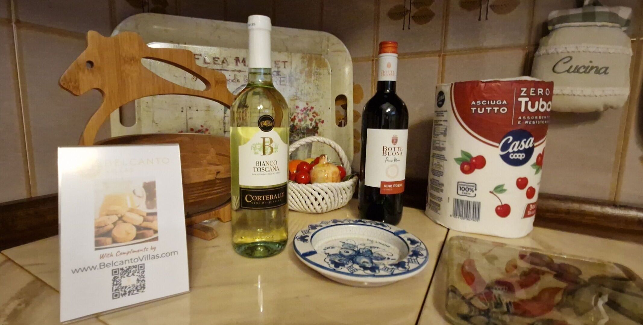 Un piano di lavoro in cucina con una bottiglia di vino bianco, una bottiglia di vino rosso, un cesto di frutta, un piatto e un vasetto di frutta in scatola.
