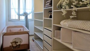 A room with a crib, a large window with curtains, and shelves with folded blankets and a vase with flowers.