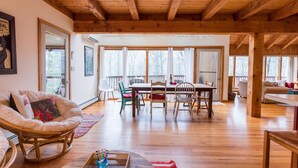 A spacious living room with a wooden ceiling, a dining area with a table and chairs, and large windows offering a view of the outdoors.