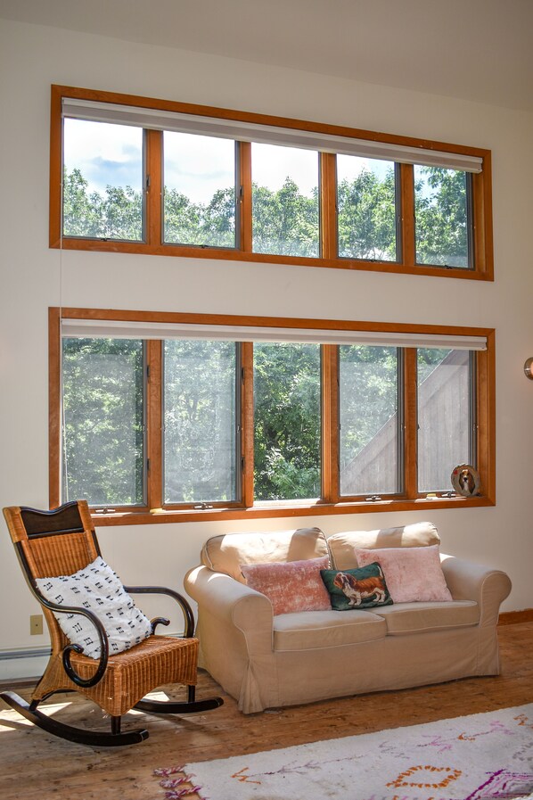 Una habitación con mecedora y sofá, cerca de ventanas amplias con vistas a árboles.