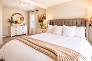 Ein Schlafzimmer mit einem großen Bett, einem Nachttisch mit Lampe, einer Kommode, einem Spiegel und einem Fenster mit Vorhängen.