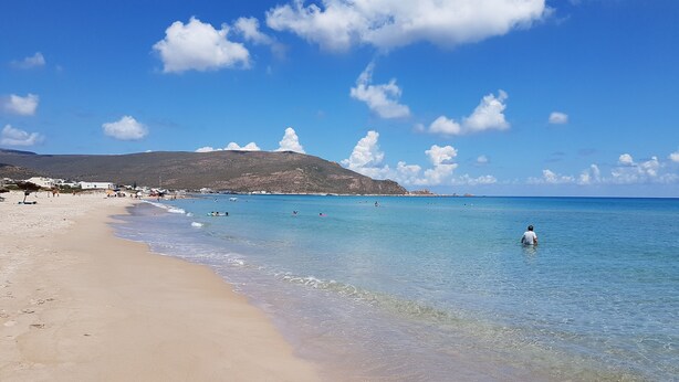 Une plage avec une eau d’un bleu limpide, un rivage sablonneux et une montagne en arrière-plan.