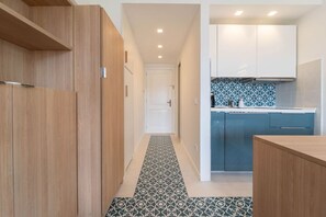 A narrow hallway with wooden wardrobes, a patterned floor, and a kitchen area with blue cabinets and patterned backsplash.
