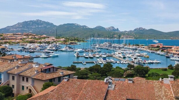 Un port de plaisance avec des bateaux amarrés, entouré de bâtiments résidentiels et avec des montagnes en arrière-plan.